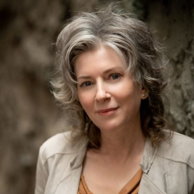 A woman with grey hair wearing a cream jacket over a rust coloured shirt.